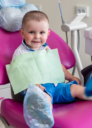 A young child sits on a pink dental chair, smiling, with a dental bib on, in a modern dentist's office, surrounded by equipment.