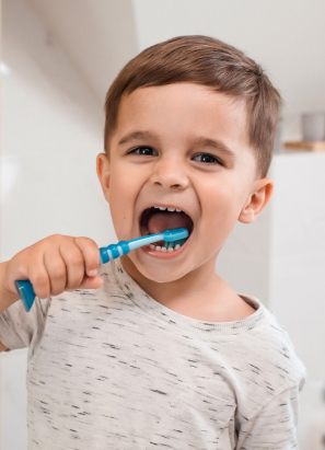 A young child is brushing their teeth with a blue toothbrush, looking directly at the camera, in a brightly lit bathroom setting.