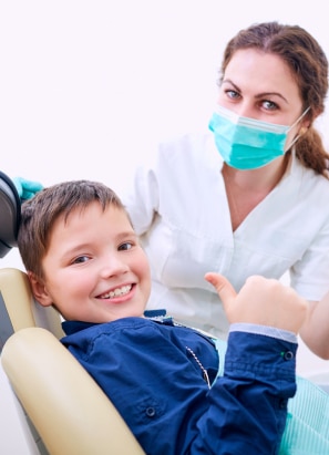 A young child smiles and gives a thumbs-up while sitting in a dental chair next to a masked person, presumably the dentist.