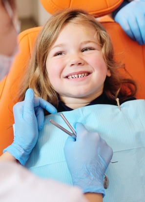 A child smiles while sitting in a dental chair, as a gloved person holding dental tools attends to them. The atmosphere appears bright and friendly.