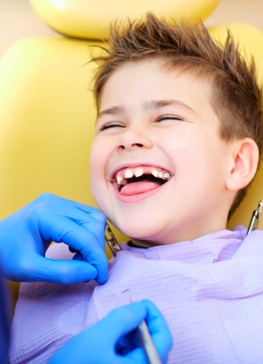 A child with missing front teeth is smiling at a dental clinic, while a person in blue gloves examines their teeth.