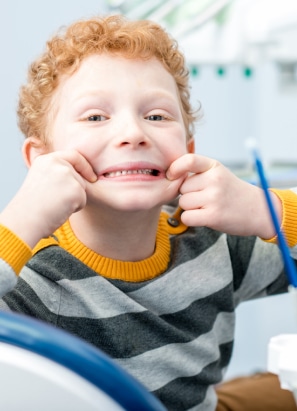 A child sits in a dental chair, pulling a playful face by stretching their lips, wearing a striped sweater with yellow accents. Dental tools are visible.