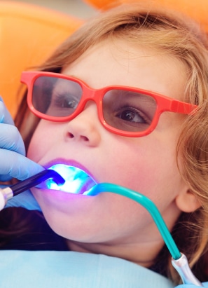 A child wearing red glasses receives dental treatment, featuring bright dental lights and tools held by a gloved person in a dental clinic.