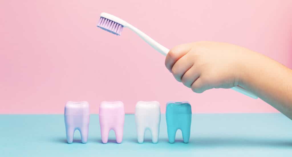 A child's hand holding a toothbrush with four colorful tooth models on a pastel blue and pink background.