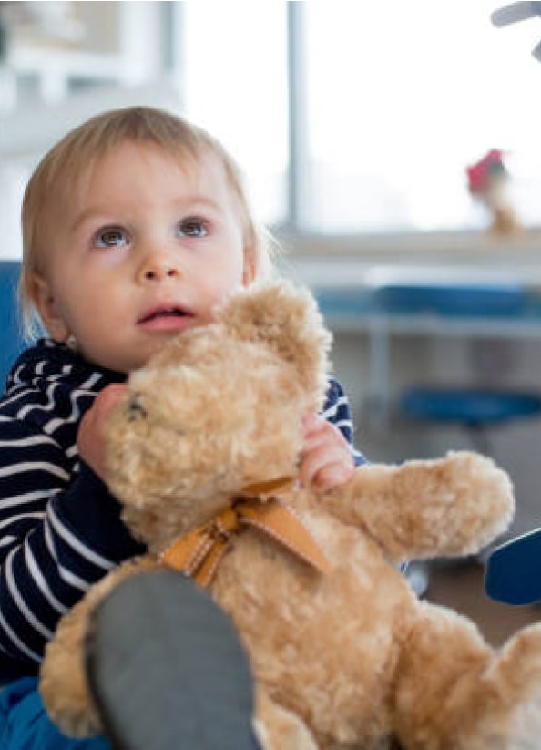 A child in a striped shirt holds a teddy bear, looking upwards. The background features blue furniture and a blurred room.