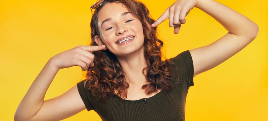 A smiling person with braces points to their teeth, standing against a solid yellow background. The person appears cheerful and confident.