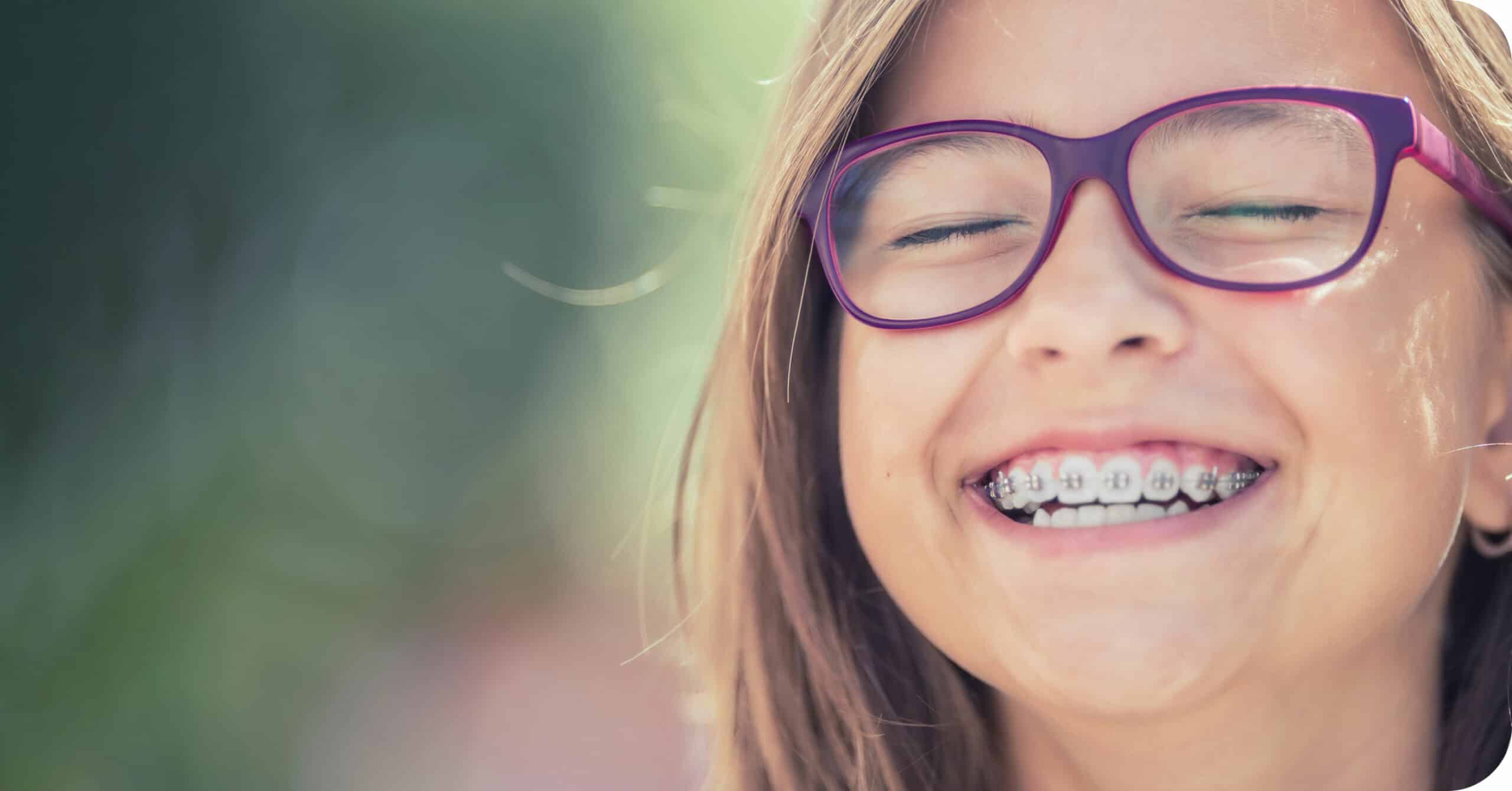 A child wearing glasses and braces smiles with closed eyes against a blurred outdoor background. No recognizable landmarks or historical buildings are visible.