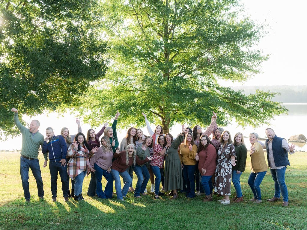 A group of people pose cheerfully in front of a large tree near a lakeside, with a bright, sunny background.