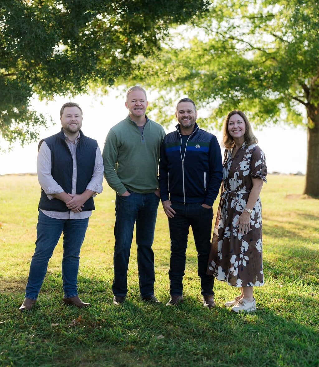 Four people standing together on grass, smiling, with trees and sunlight in the background. The setting is casual and outdoors.