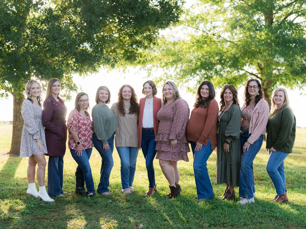 A group of ten people stand on grass in front of large trees, with sunlight filtering through the leaves.