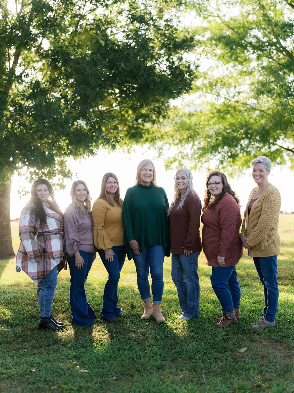 Seven people stand outdoors on a grassy field, with trees in the background. They wear colorful sweaters and smile at the camera.