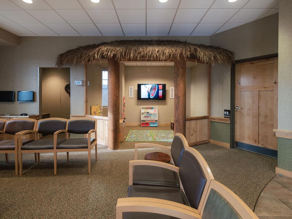 A waiting room features wooden chairs, television, children's play area, and tropical-themed decor with a thatched roof accenting the entrance.