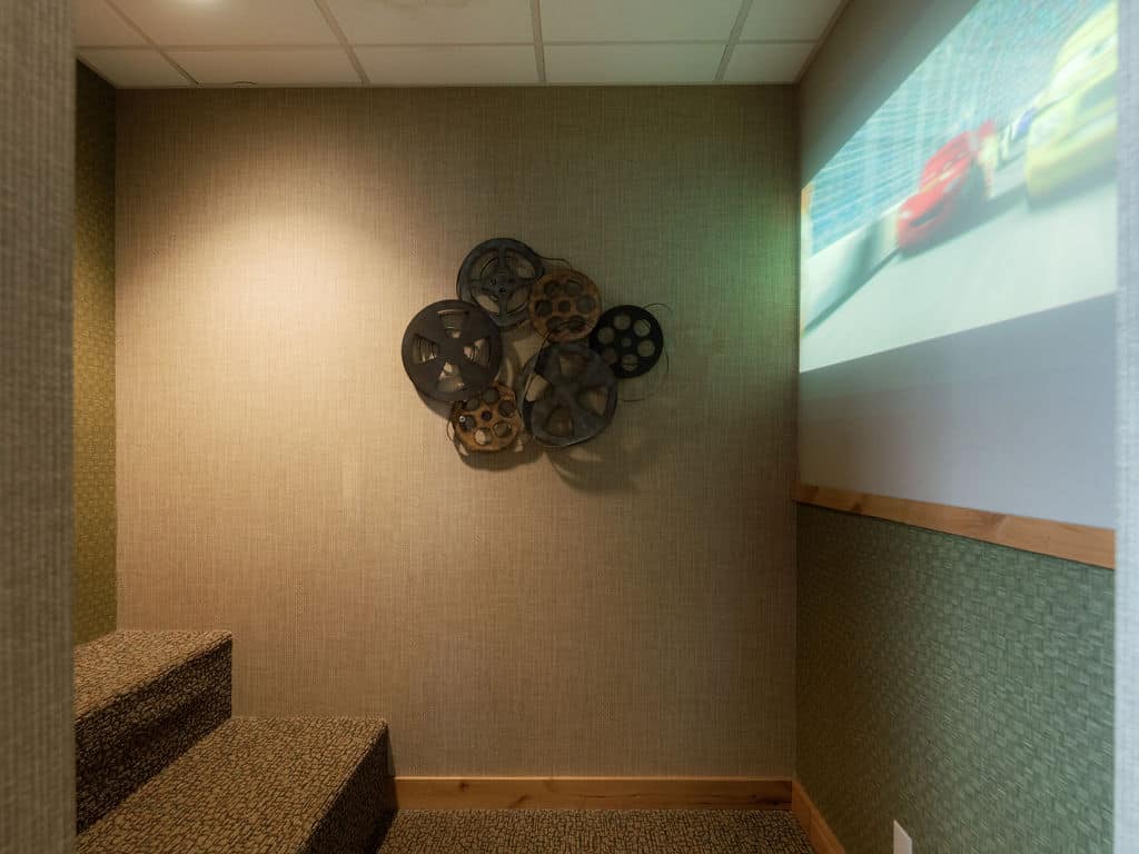 Home theater corner with film reel wall decor. A movie is projected on the screen, steps with textured carpet lead up on the left.