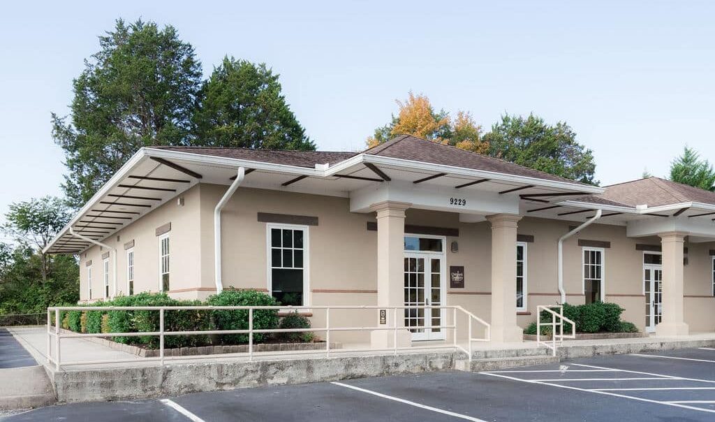 A single-story beige building with large windows, surrounded by trees, and an empty parking lot. The roof has extended eaves and white trim.