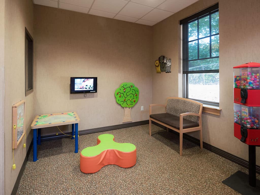 A small waiting room with children's play area, featuring a wall-mounted TV, toys, a bench, and a gumball machine by the window.