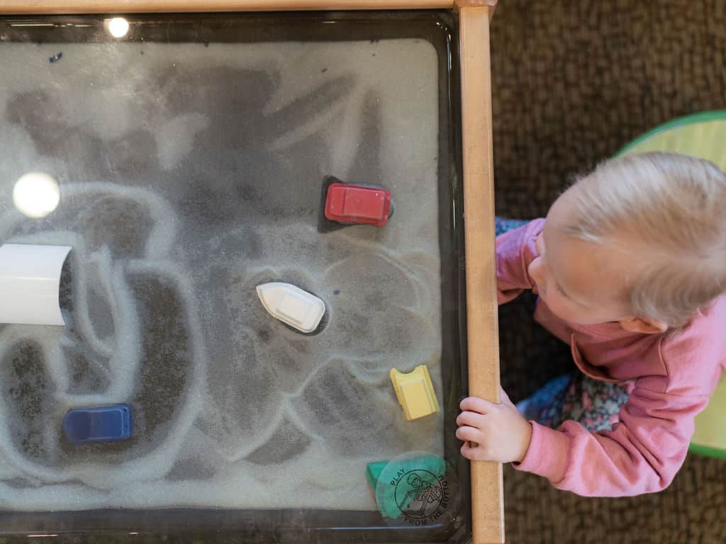 A child is playing with toy vehicles on a sand table, observing their movements. The setting appears to be indoors.