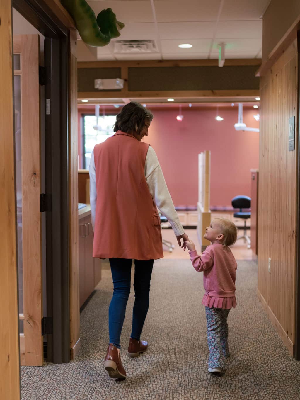 A person and child walk hand-in-hand through a warmly lit hallway with wooden decor and visible office furniture in the background.