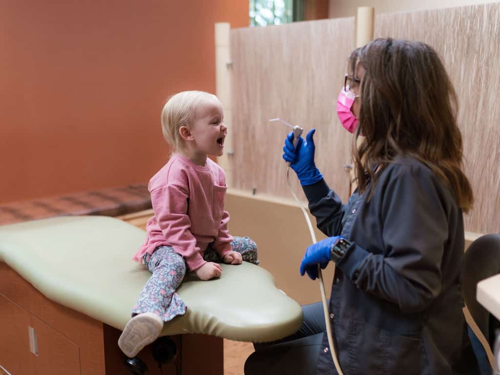 A child sits on a dental chair, mouth open, as a person wearing gloves and a mask prepares to examine them, creating a caring atmosphere.