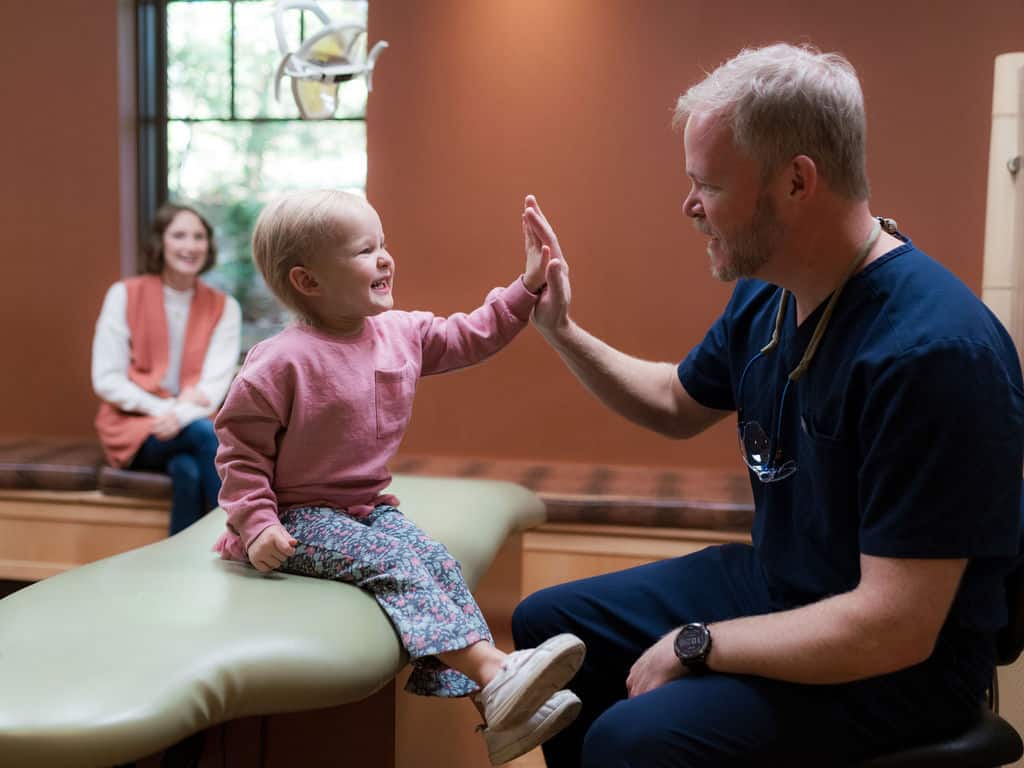 A child high-fives a seated person in a dental office, while another person watches from a background bench near a window.