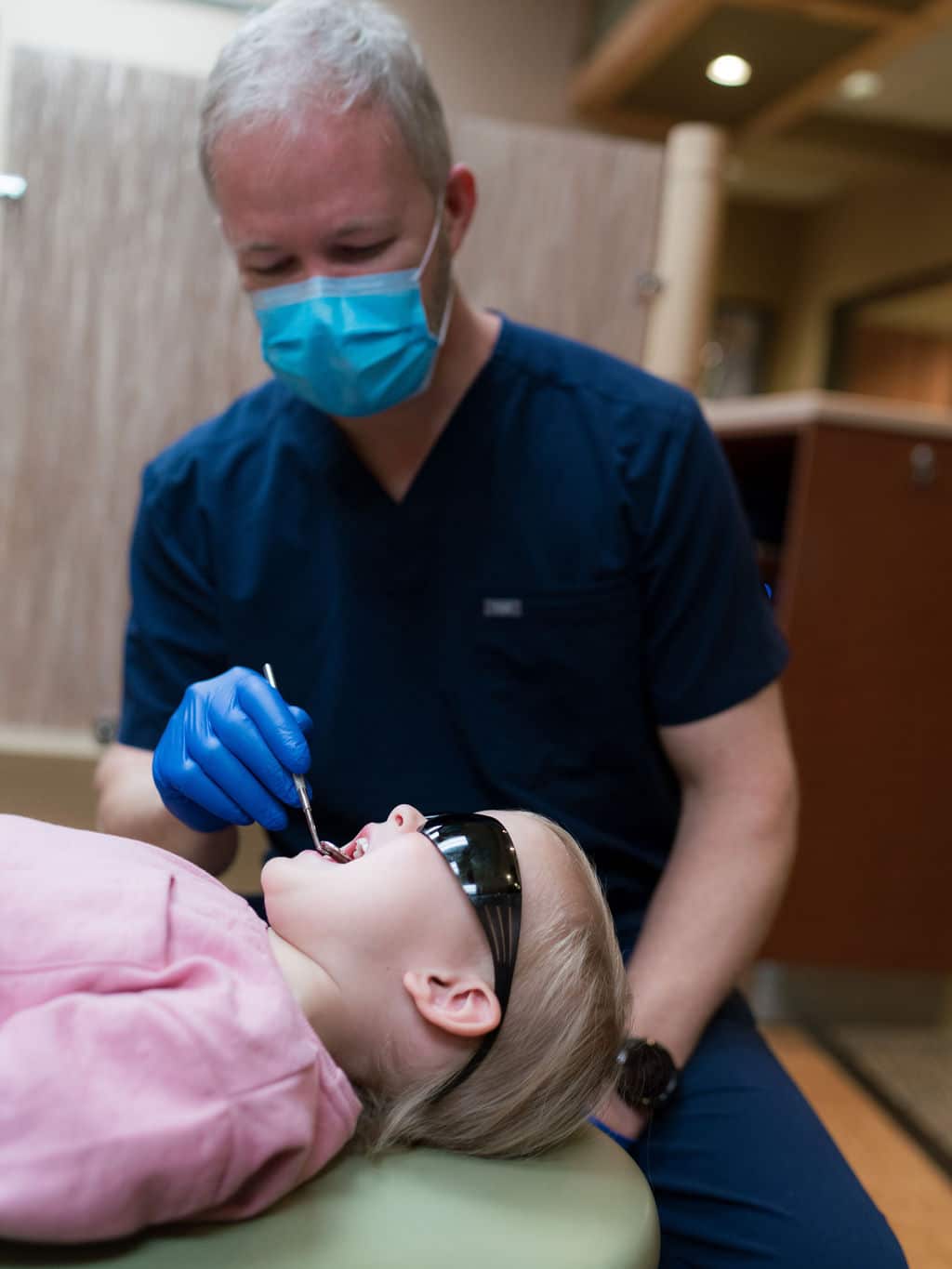 A person wearing medical scrubs and mask examines a child’s teeth in a dental office, using tools. The child wears sunglasses.