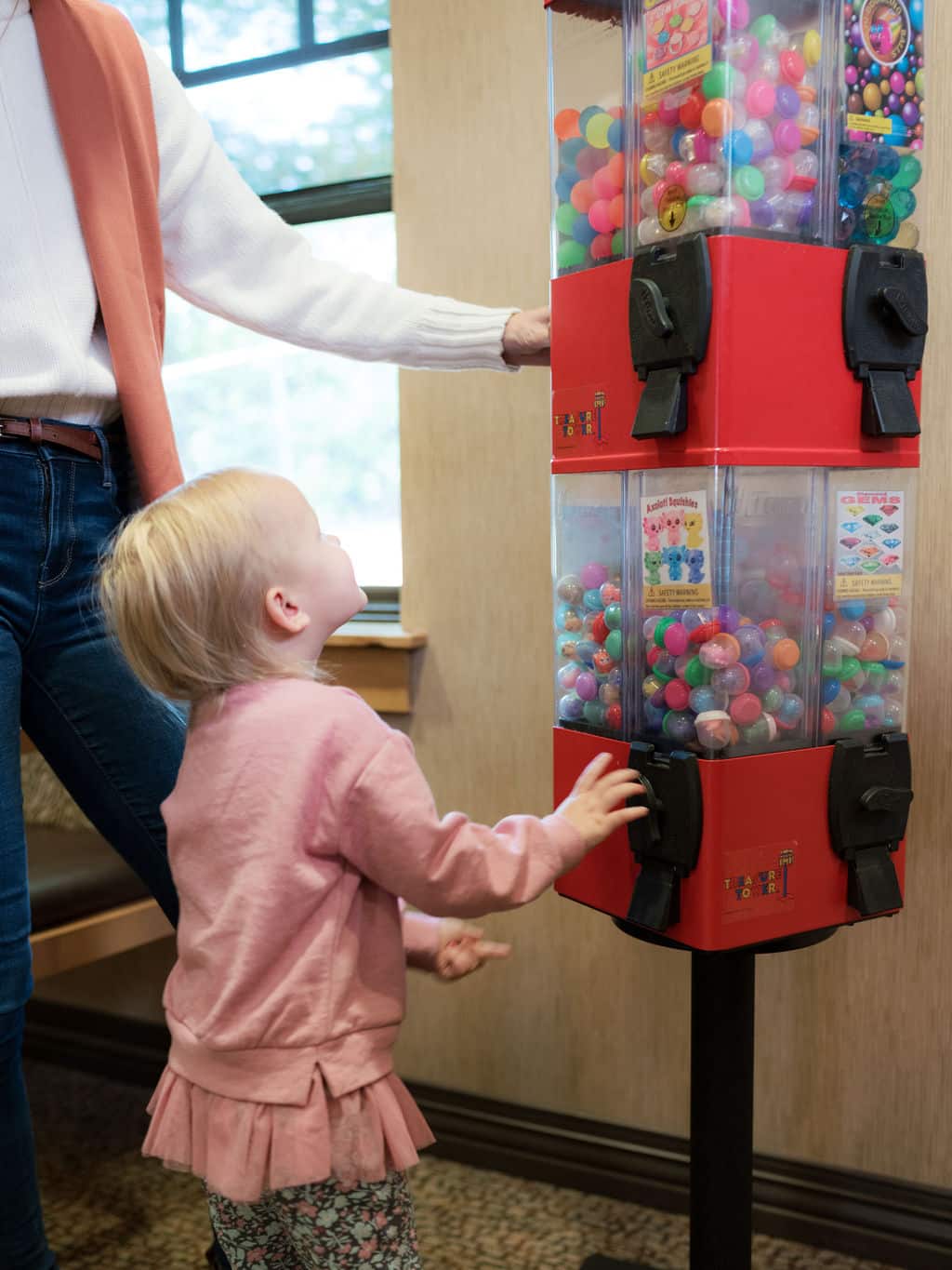 A child and person interact with a red gumball vending machine indoors, next to a window. The machine is filled with colorful gumballs.