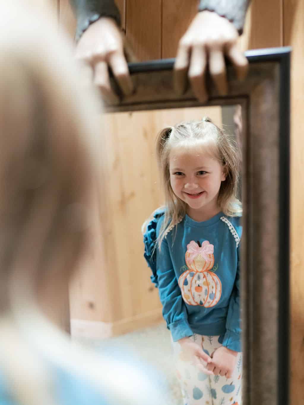 A child, wearing a pumpkin-themed shirt, smiles in front of a mirror with a frame held by unseen person’s hands in a wooden room.