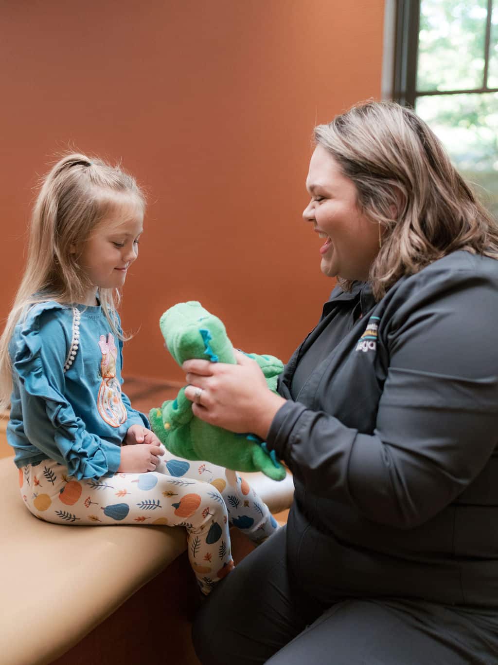 A person and child share a joyful moment indoors, playing with a green stuffed animal, against a brown wall and large window.