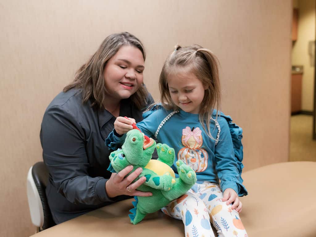 A person and a child interact with a dinosaur toy, teaching dental hygiene in a bright, professional setting.