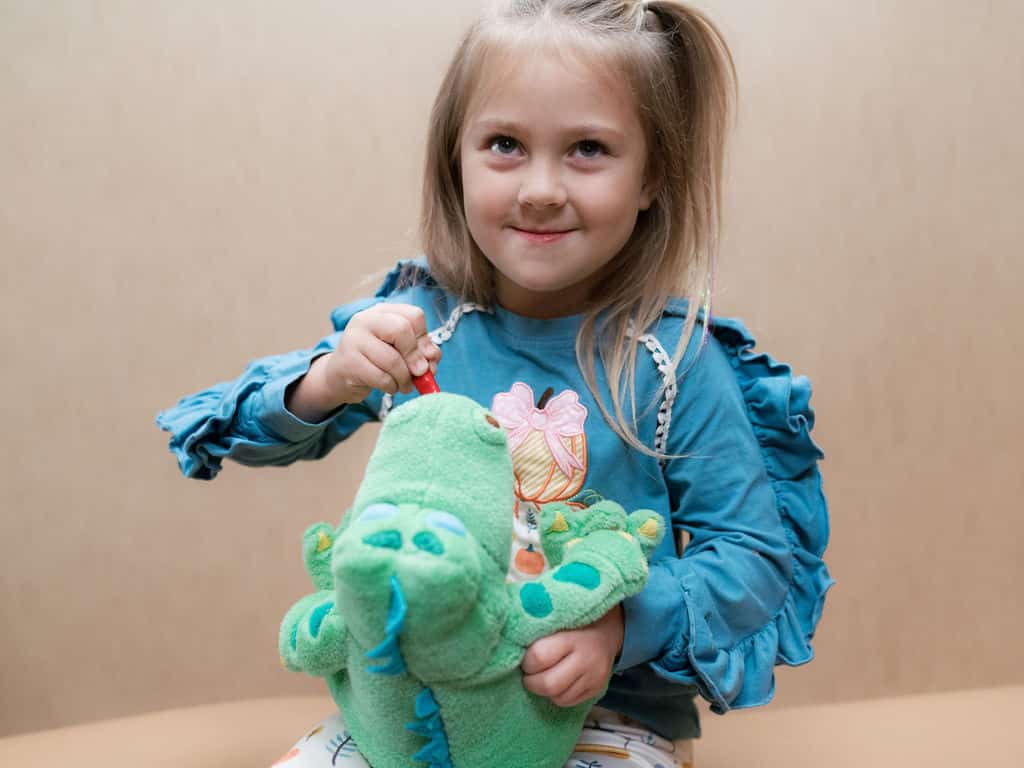 A child smiles while playfully using a toothbrush on a green dinosaur toy, sitting against a simple beige background.