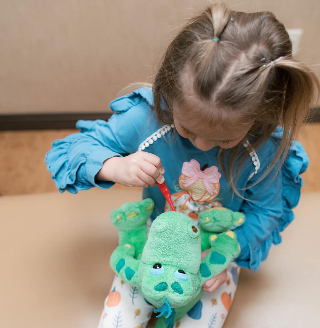 A child in colorful clothes plays with a green dinosaur toy, sitting on a bench, focusing on interaction in an indoor setting.