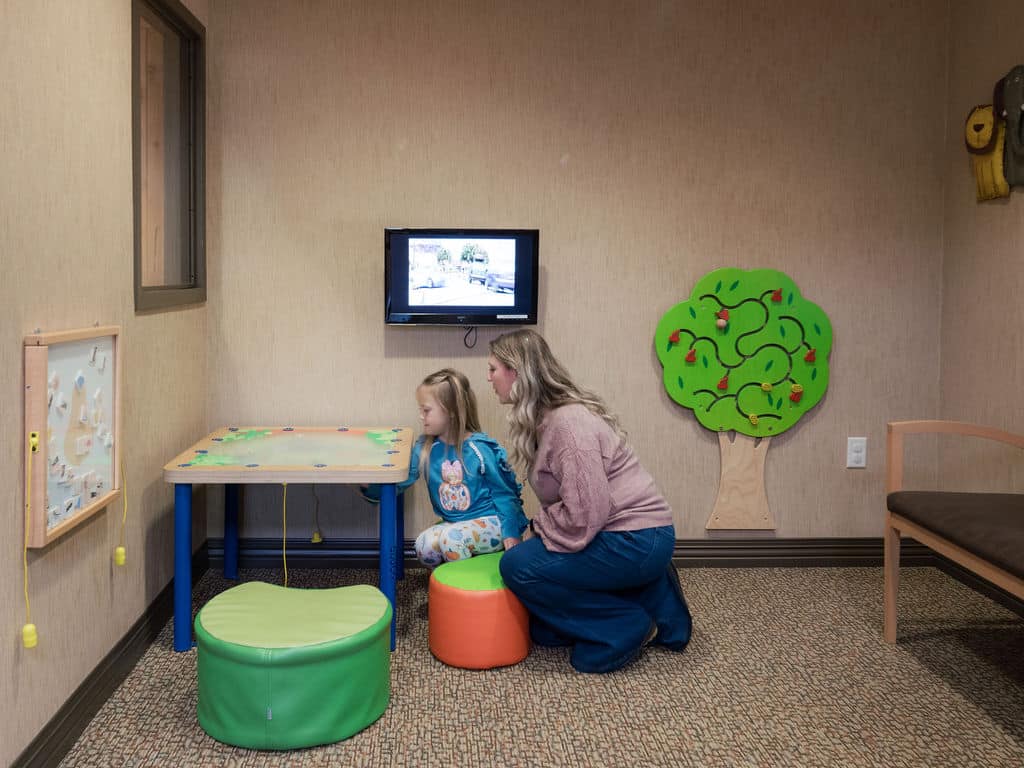 A child and person sit at a small table in a room with a TV and colorful decor, including a wall-mounted tree design.