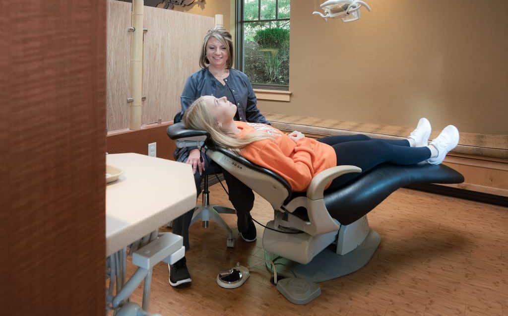 A person reclines in a dental chair, another person standing nearby, with a monitor overhead in a warmly decorated room.