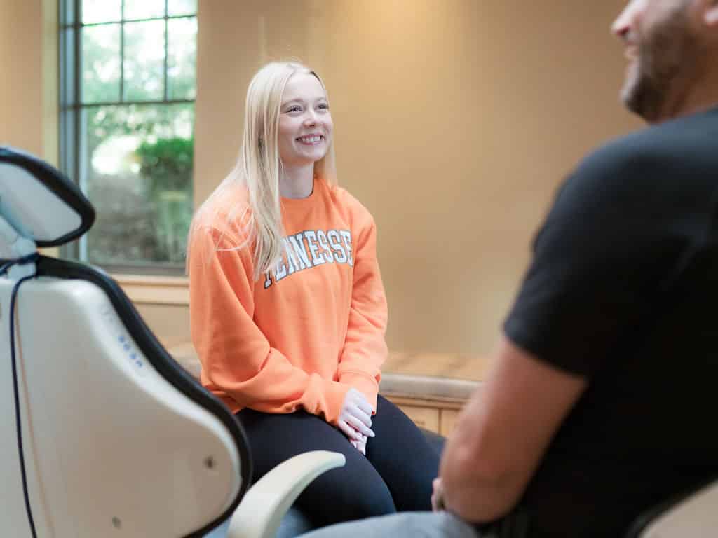 A person in an orange Tennessee sweatshirt smiles, sitting in a room with a large window beside another person.