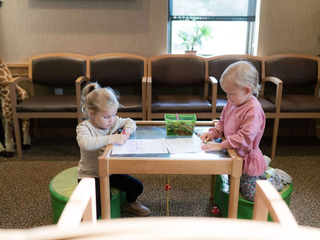 Two children are drawing at a small table in a cozy room with wooden chairs and carpet. A stuffed giraffe stands nearby.