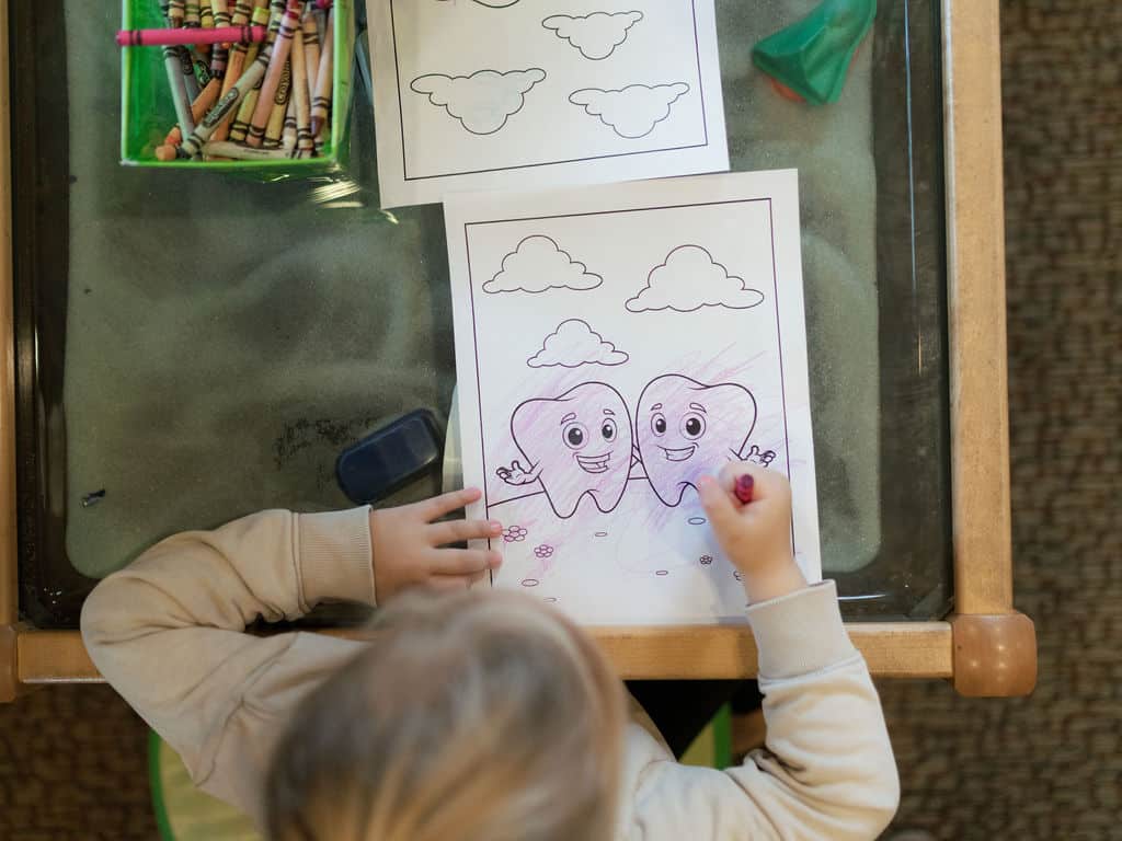 A child is coloring a drawing of two smiling teeth on paper at a table with crayons and an eraser.