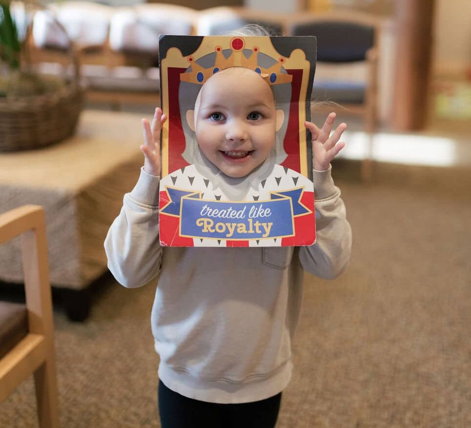 A child holds a whimsical cutout reading "treated like royalty" in a cozy room with soft furnishings and a potted plant.