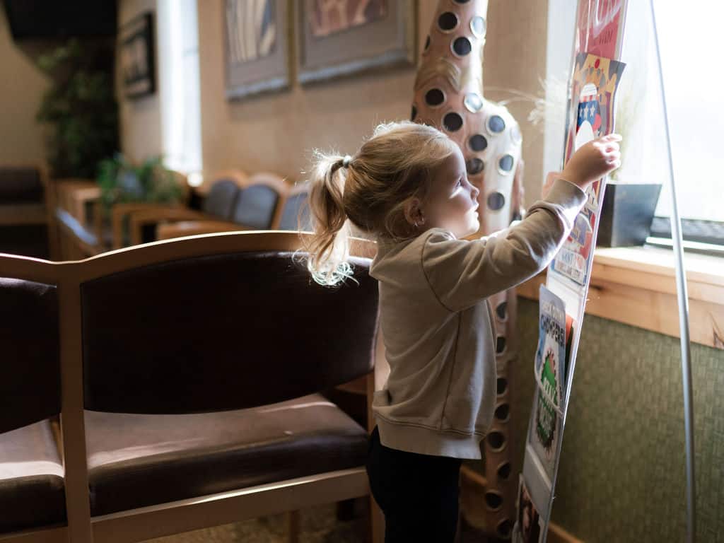 A child in a cozy, sunlit room interacts with a stand displaying colorful pictures. Chairs and framed artwork decorate the background.