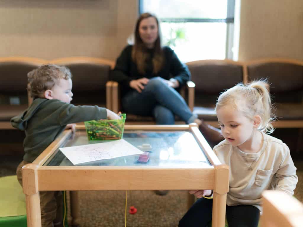 Two children play with toys at a table in a waiting area while a person sits on a chair in the background.