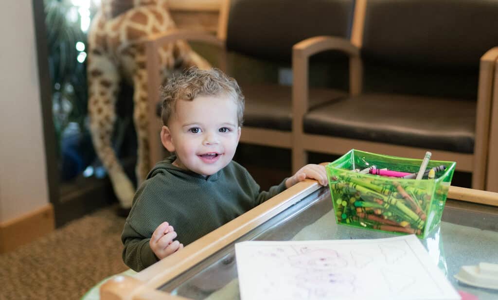 A child smiles at a small table with crayons and papers; a stuffed giraffe stands nearby in a cozy room.