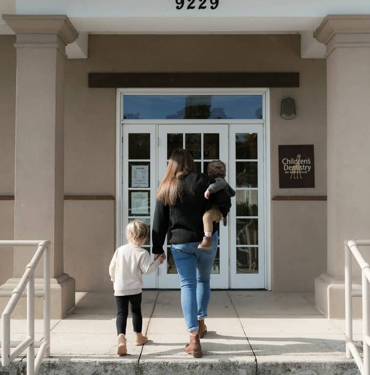 A person with two children approaches the entrance of a children's dentistry clinic. The building appears modern with beige pillars and a glass door.