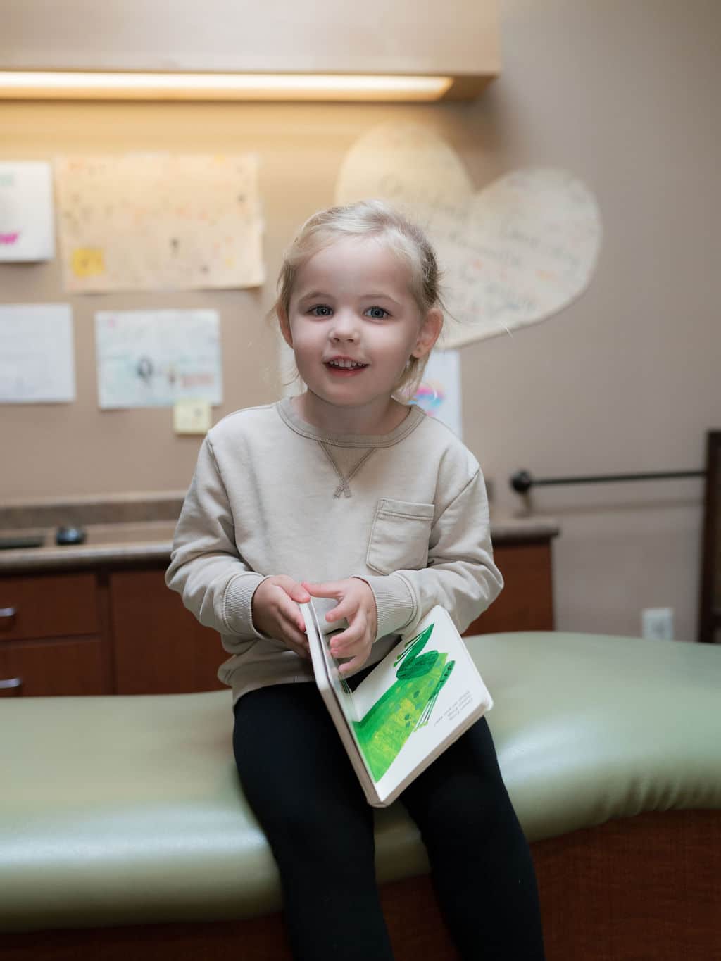 A child sits on an examination table, holding a book. The background features various children's drawings and colorful art on the wall.