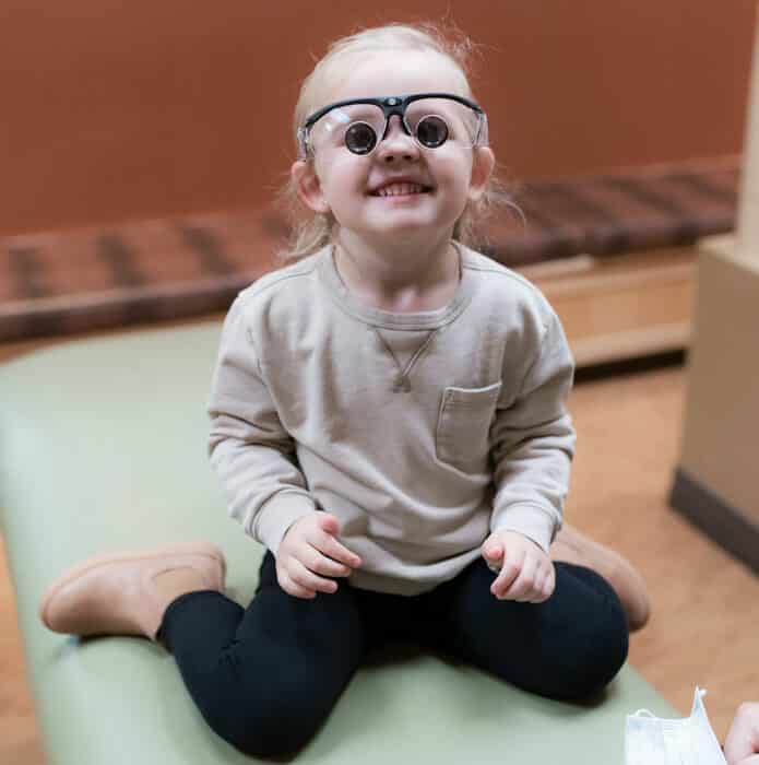 A child with glasses is smiling while sitting on an examination table, with a person holding a mask nearby in a clinic room.