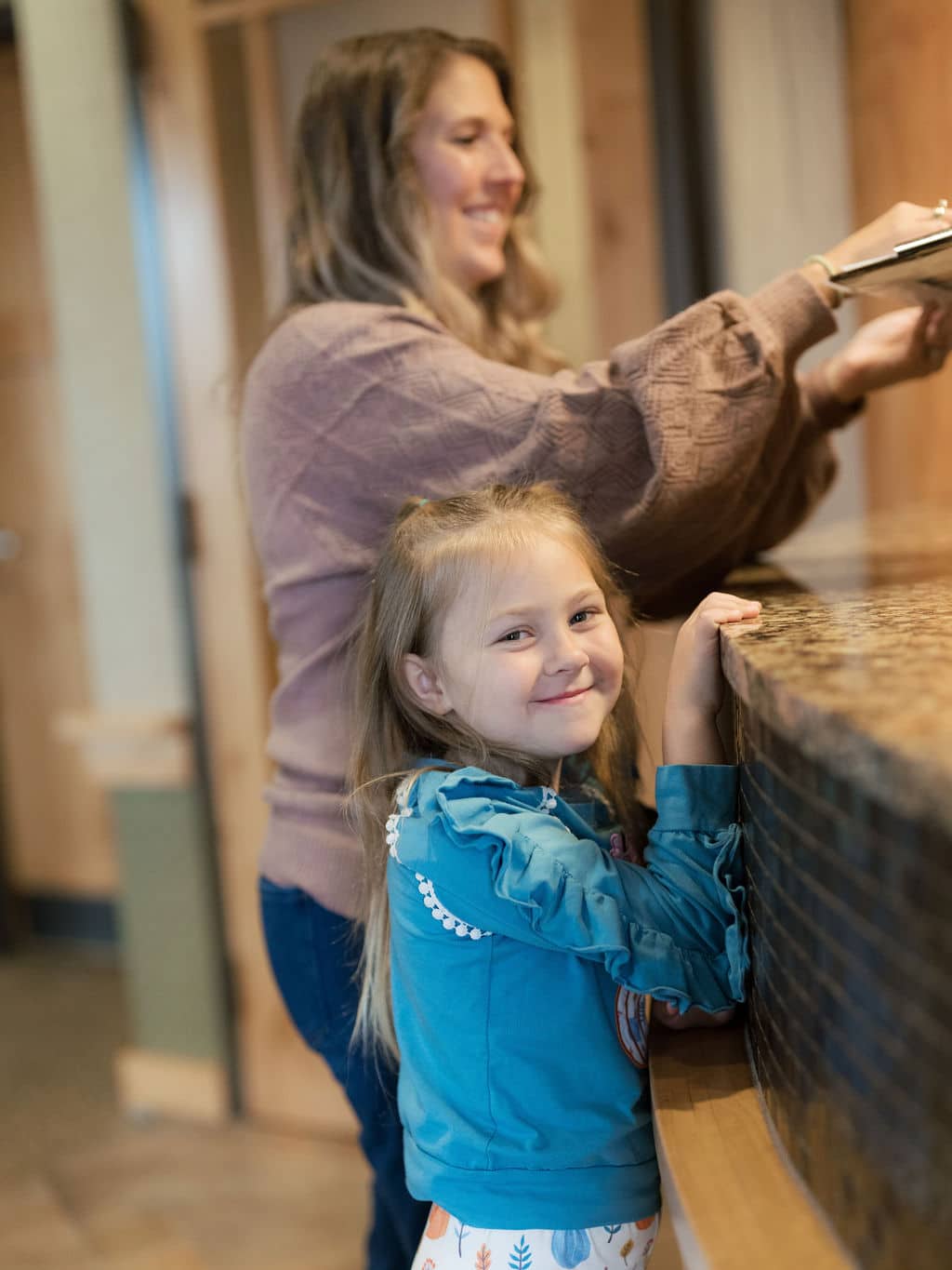 A person and a smiling child stand at a textured counter in an indoor setting, with wooden and neutral-colored walls.