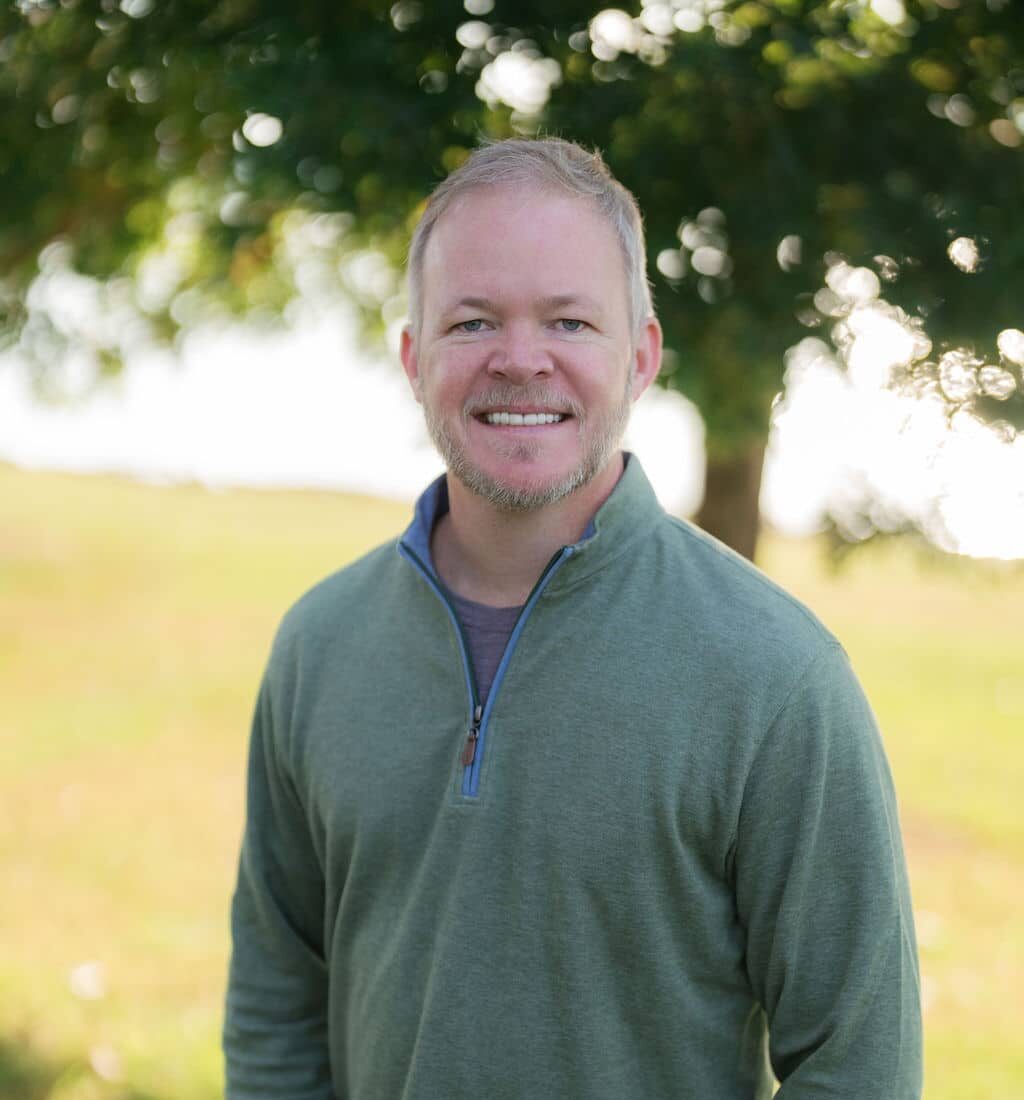 A person in a green sweater stands smiling outdoors with trees and a blurred grassy field in the background on a sunny day.