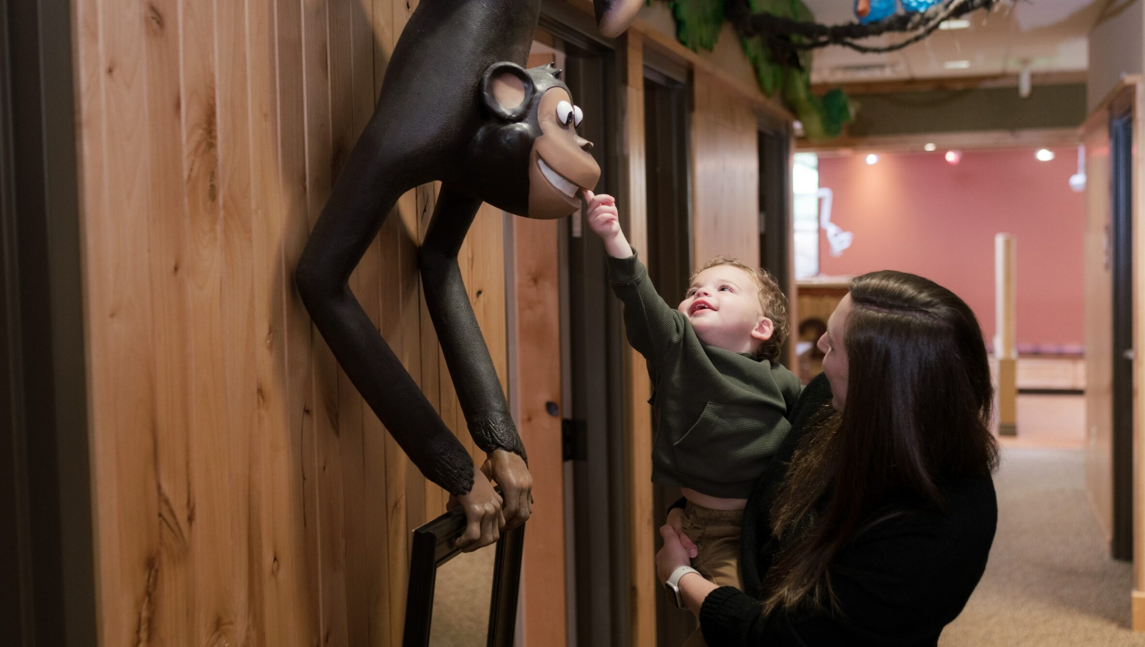 A person holds a child reaching for a monkey sculpture in a warmly lit interior space with wood-paneled walls and overhead greenery.