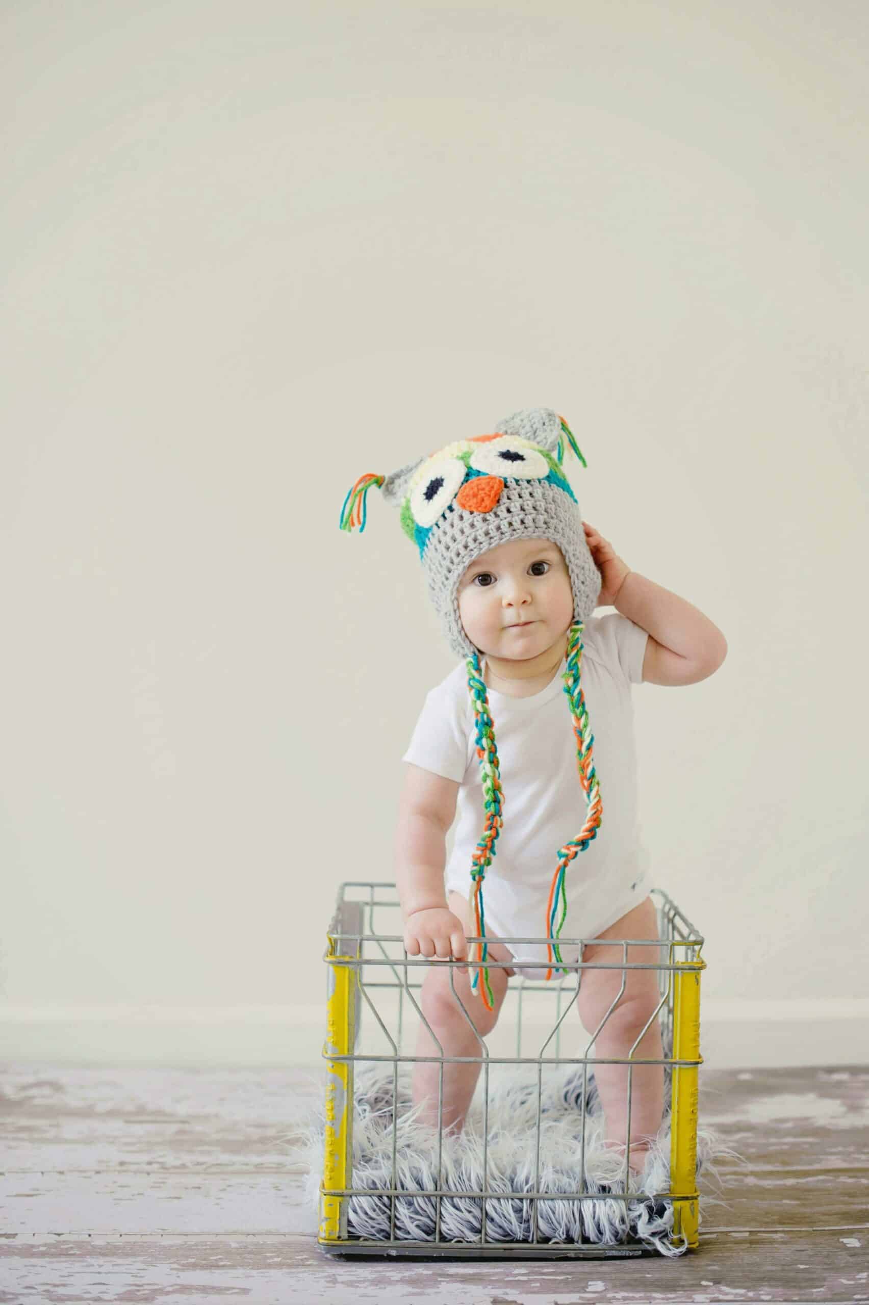 A baby wearing an owl-themed knit hat sits inside a metal basket on a wooden floor, against a plain background.