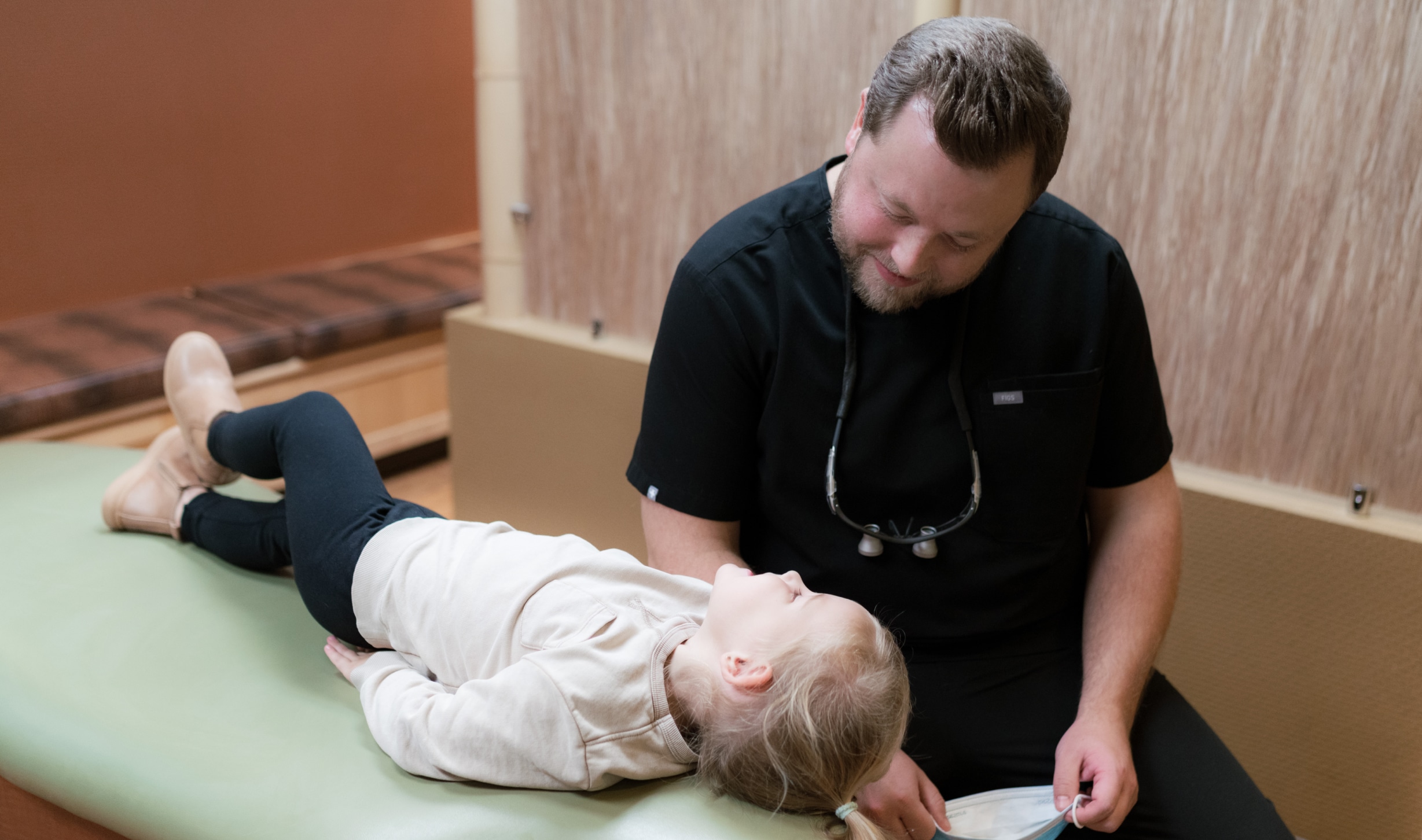 A person in dental attire interacts kindly with a child lying on an examination table in a dental office setting.