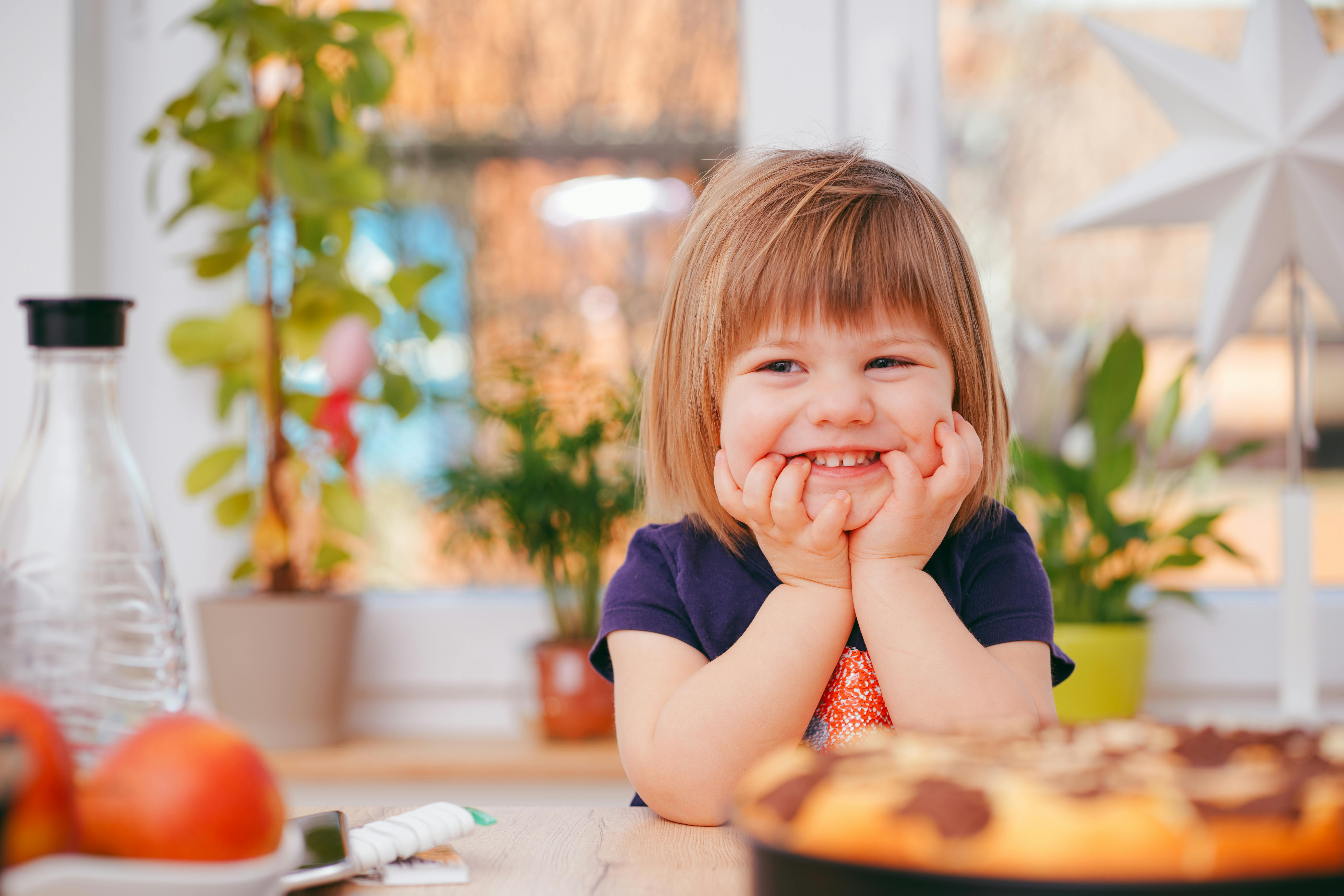 A smiling child in a festive room with plants, a star decoration, fruit, cake, and a bottle on a table.