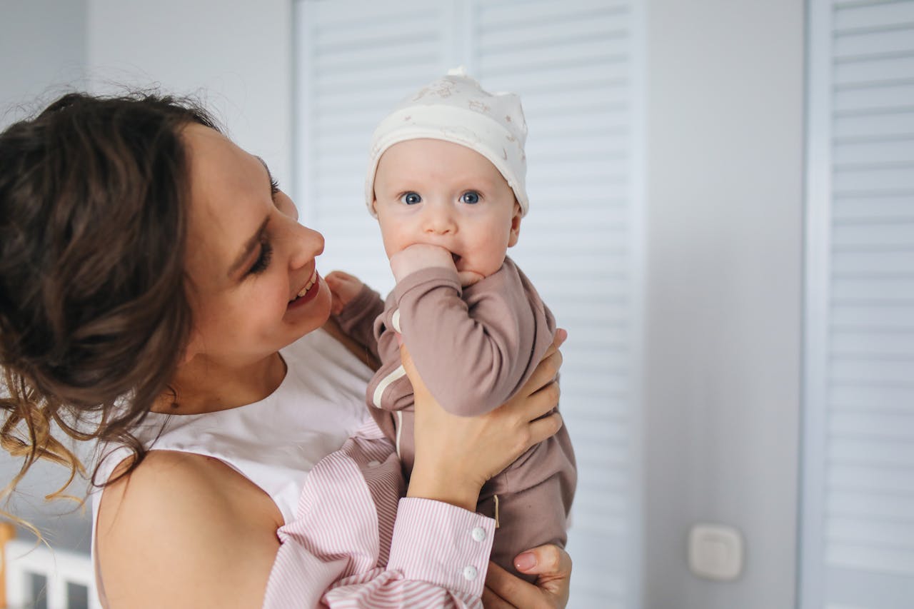 A person holding a baby smiles warmly. The baby, wearing a hat, looks curious. White shutters are in the background, creating a cozy atmosphere.