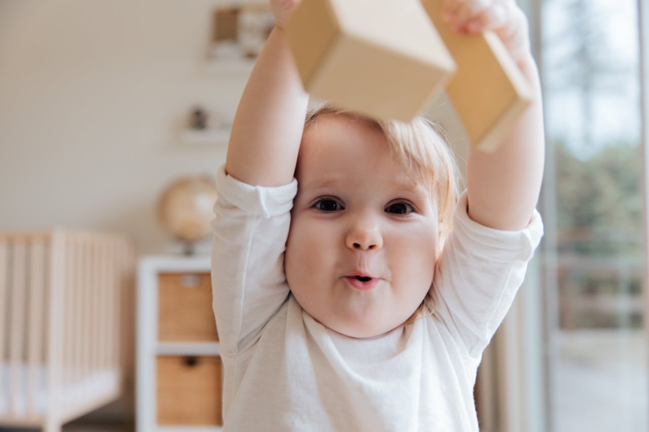 A child joyfully holds up wooden blocks in a cozy room with a crib and soft lighting, expressing excitement and playfulness.